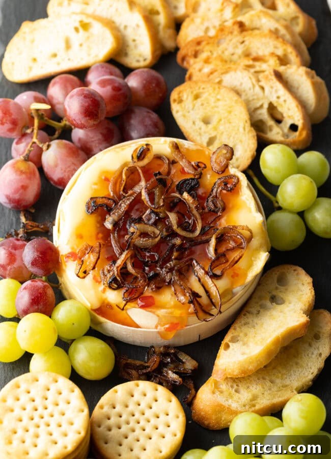 Gooey Baked Camembert Delight 12 Wheel of Camembert cheese baked with sweet chili sauce and sauteed shallots. It is on a platter and surrounded by red and green grapes, crackers, and baguette slices.
