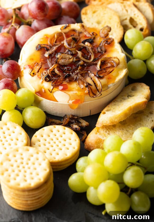 Gooey Baked Camembert Delight 13 Wheel of Camembert cheese baked with sweet chili sauce and sauteed shallots. It is on a platter and surrounded by red and green grapes, crackers, and baguette slices.