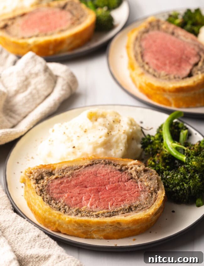 Slice of beef wellington on a plate with mashed potatoes and broccolini.