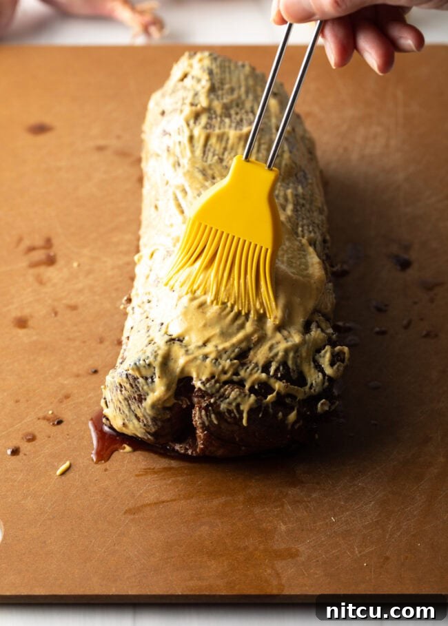 Hand with yellow basting brush, brushing Dijon mustard over the seared tenderloin.