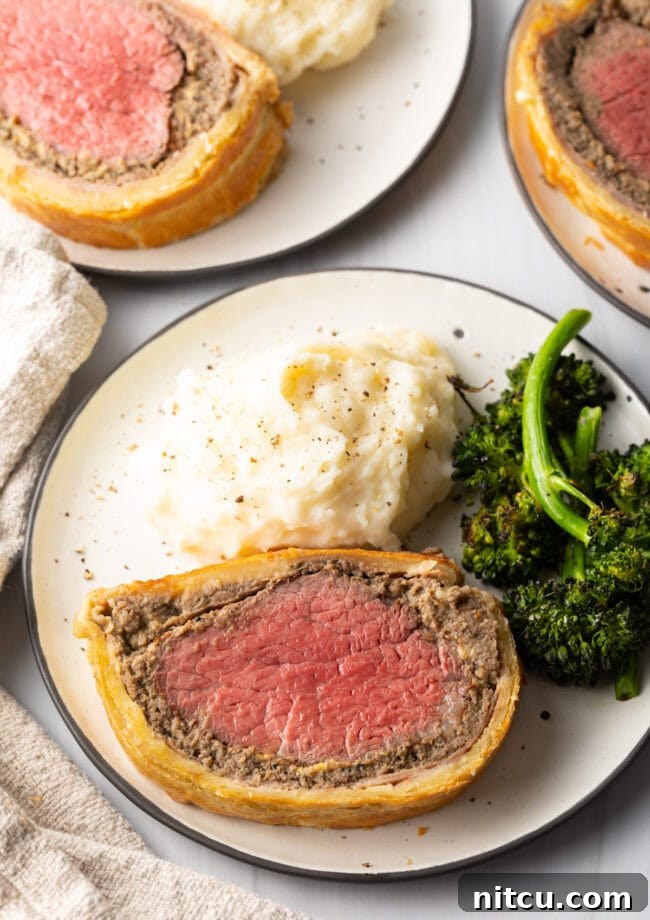 Slice of wellington on a plate, with broccoli and mashed potatoes.