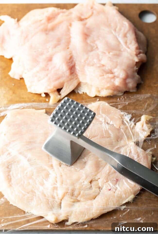 Savory Stuffed Chicken Rolls 5 Butterflied chicken breasts on a cutting board, covered in plastic and with a metal mallet pounding them thin.