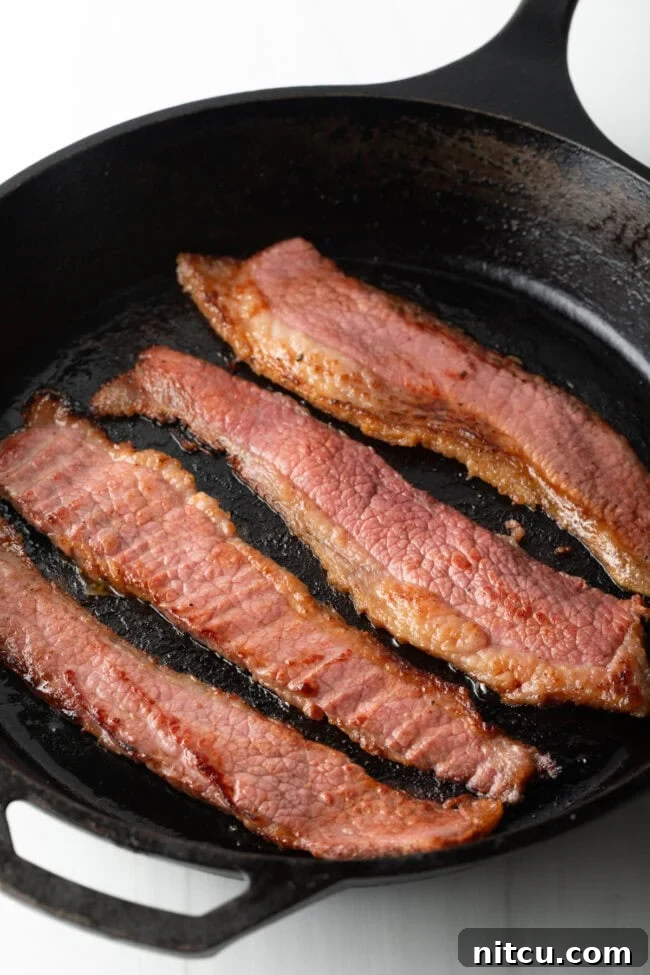 Perfect Homemade Beef Bacon 2 Four strips of beef bacon cooking in a cast iron skillet.
