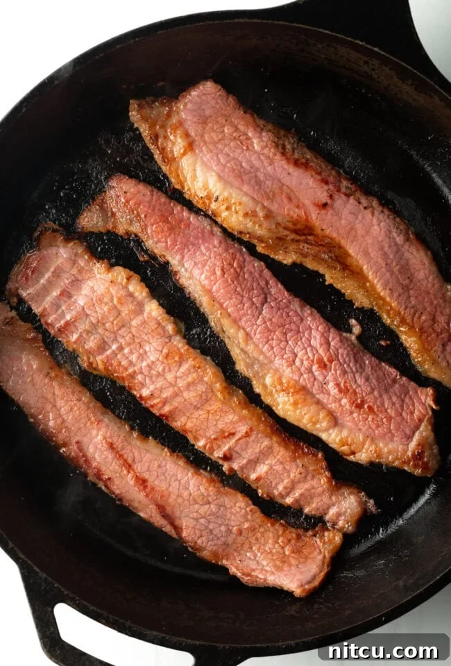 Perfect Homemade Beef Bacon 3 Top down view 4 pieces of bacon made with beef cooking in a cast iron skillet.