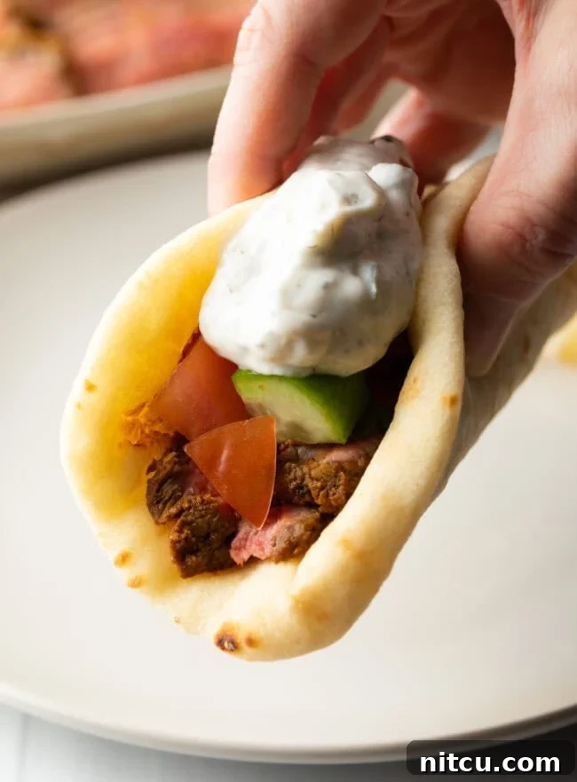 Authentic Street Style Beef Shawarma 14 Close view hand holding flatbread naan beef shawarma recipe topped with chopped cucumbers, tomatoes, red onion and tzatziki sauce.
