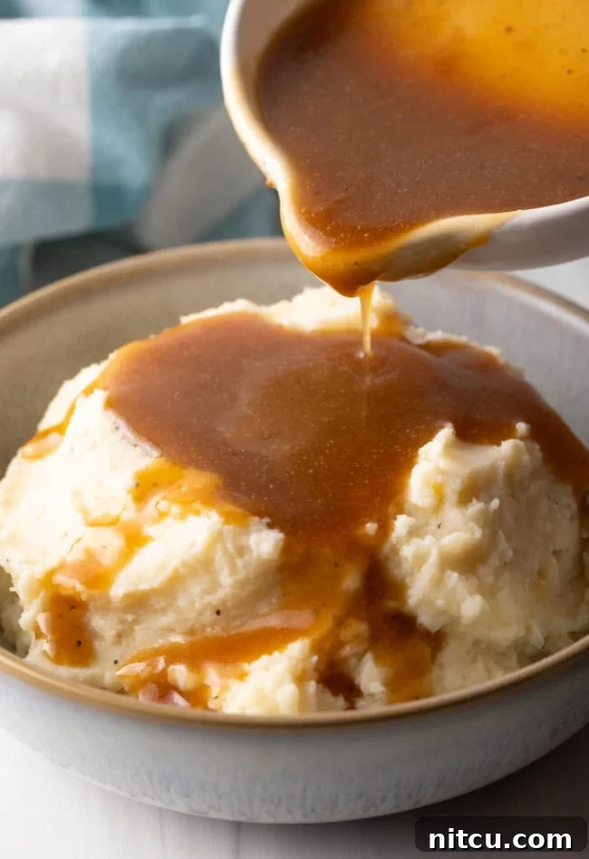 Pouring homemade beef gravy from a white pitcher over a bowl of mashed potatoes, showcasing its smooth texture.