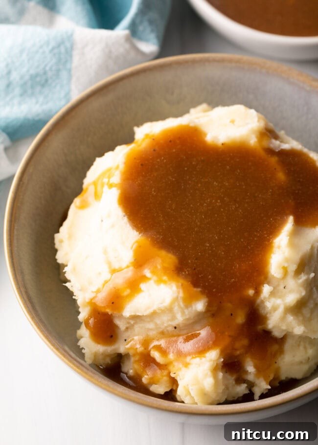 A large bowl of mashed potatoes covered with a generous amount of homemade beef gravy, with a white pitcher in the background.