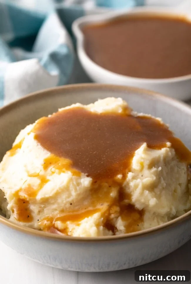 A beautiful bowl of mashed potatoes generously covered with homemade beef gravy, with a white pitcher of more gravy in the background, ready to be served.