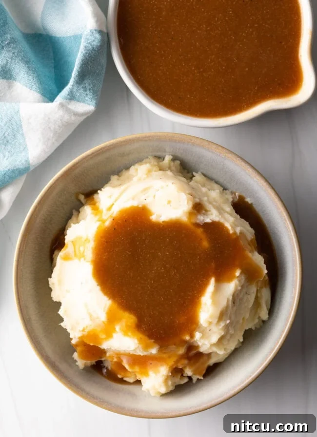 Top down view of a bowl of creamy mashed potatoes generously covered with homemade beef gravy, with a white pitcher of more gravy in the background.