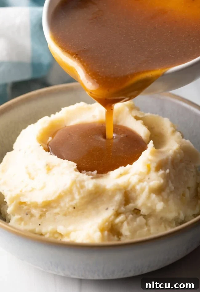 Pouring homemade beef gravy from a white pitcher over a bowl of mashed potatoes, highlighting its rich, pourable texture.
