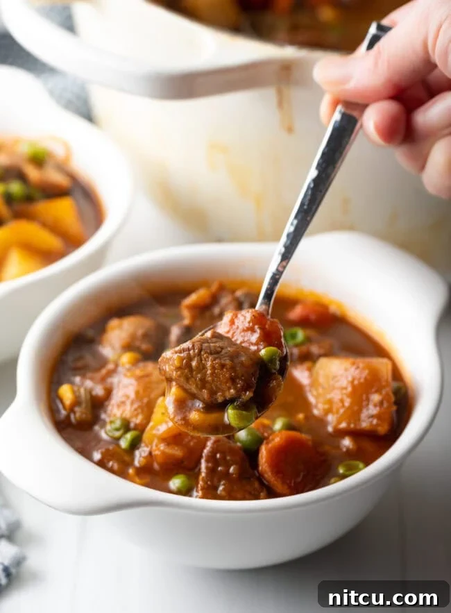 Kitchen Sink Stew 2 Mulligan beef stew with chopped potatoes, carrots, peas, and chunks of meat in a white bowl. Hand with metal spoon showing a spoonful to camera.