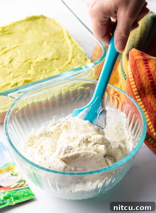 Fiesta Bean Stack 7 Hand with blue spatula, blending sour cream layer ingredients together in a small glass bowl.