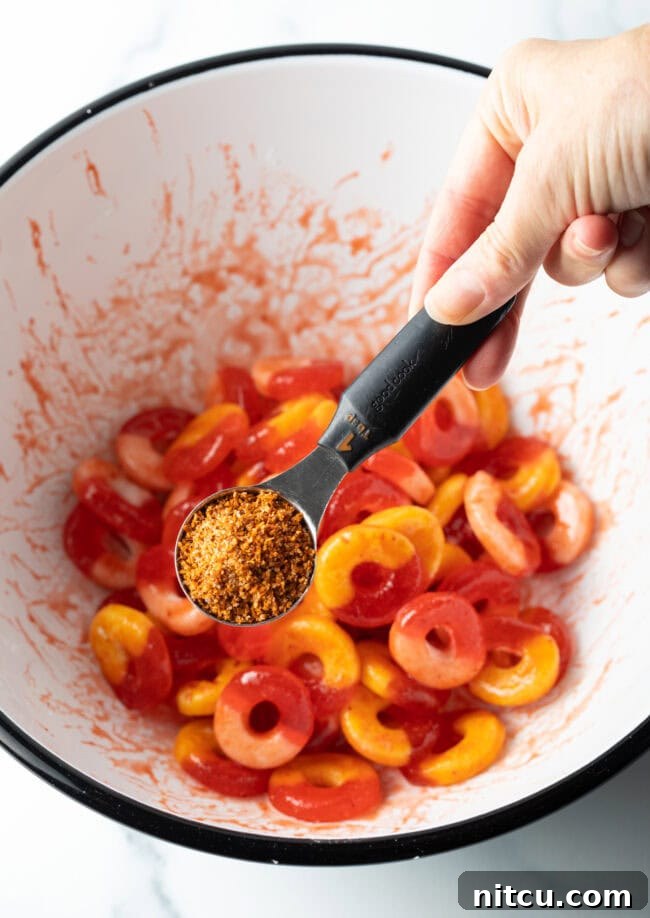Spicy Chamoy Mexican Candies 6 Hand adding Tajin seasoning from a metal measuring spoon into a bowl of gummy rings coated in chamoy.