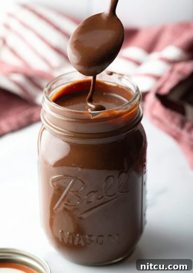Metal spoon pouring the hot fudge sauce into a glass mason jar, showcasing its rich, thick consistency.