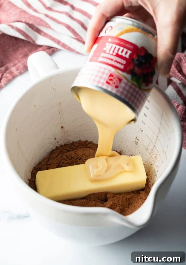 A hand adding sweetened condensed milk to a white bowl containing sifted cocoa powder and butter cubes.