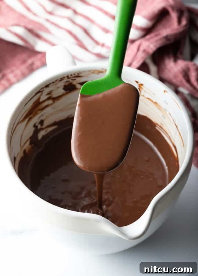 A green spatula held above a white bowl filled with freshly made homemade hot fudge sauce.