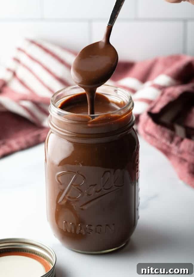 Metal spoon pouring rich homemade chocolate fudge sauce into a clear glass mason jar for storage.