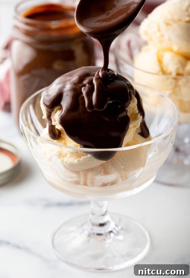 A spoon expertly drizzling homemade hot fudge sauce over a glass bowl of creamy vanilla ice cream, creating a tempting swirl.