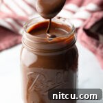 Metal spoon pouring homemade chocolate fudge sauce into a glass mason jar.