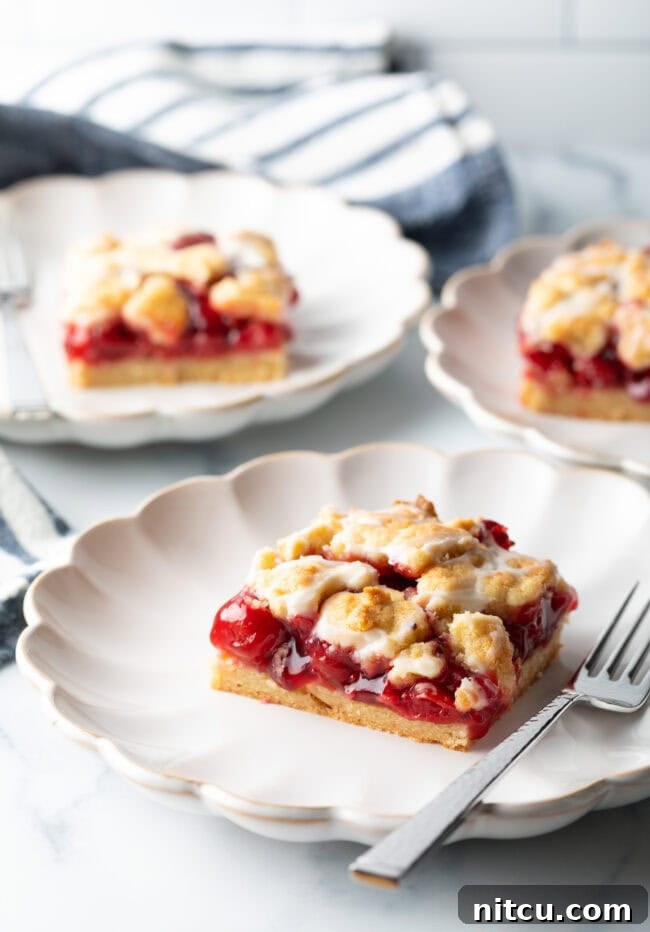 Sweet Cherry Crumble Bars 13 Three individual pieces of cherry pie bars, glistening with a sugar glaze, artfully arranged on white scalloped plates.
