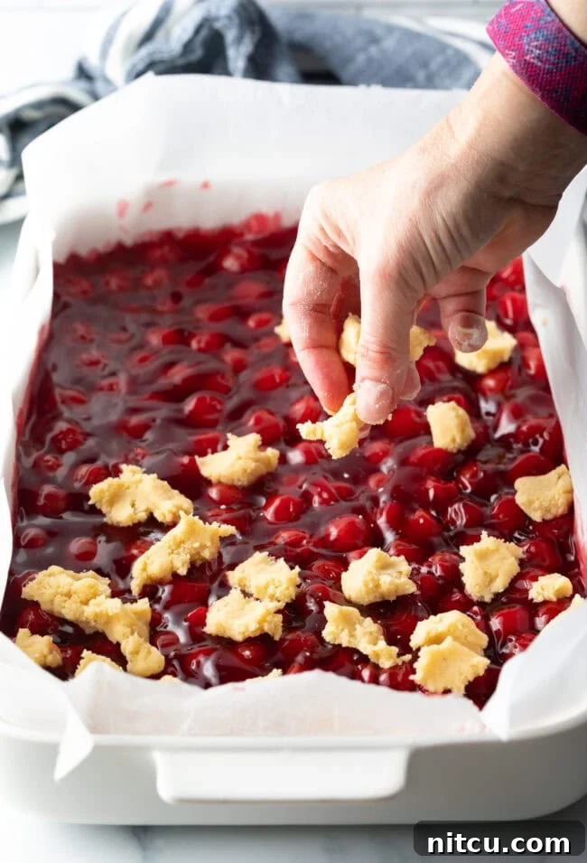 Sweet Cherry Crumble Bars 9 A hand carefully adds small, raw dough pieces onto the surface of the cherry pie filling, creating a rustic topping for the bars.