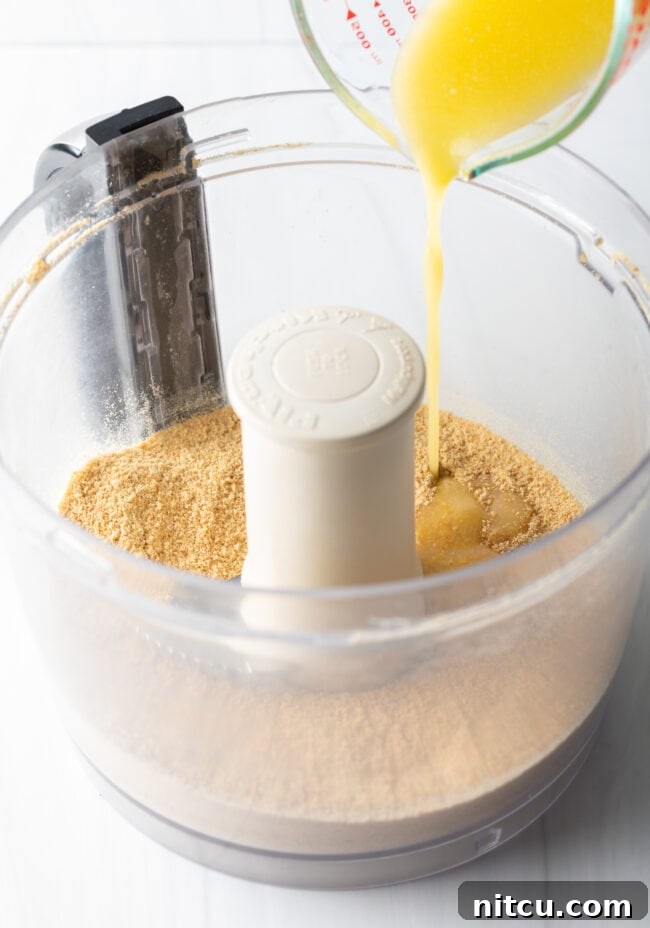 Melted butter being poured over crushed graham crackers in a food processor, initiating the crust-making process.