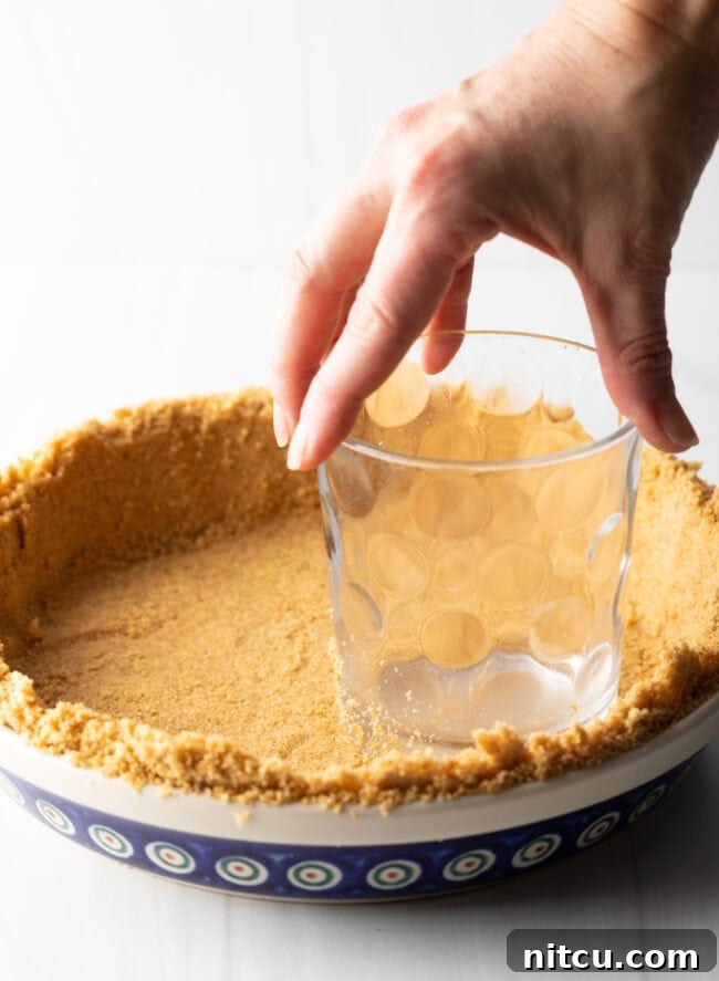 A hand using a clear glass to firmly press graham cracker crumbs into a pie plate, forming an even and compact pie crust.