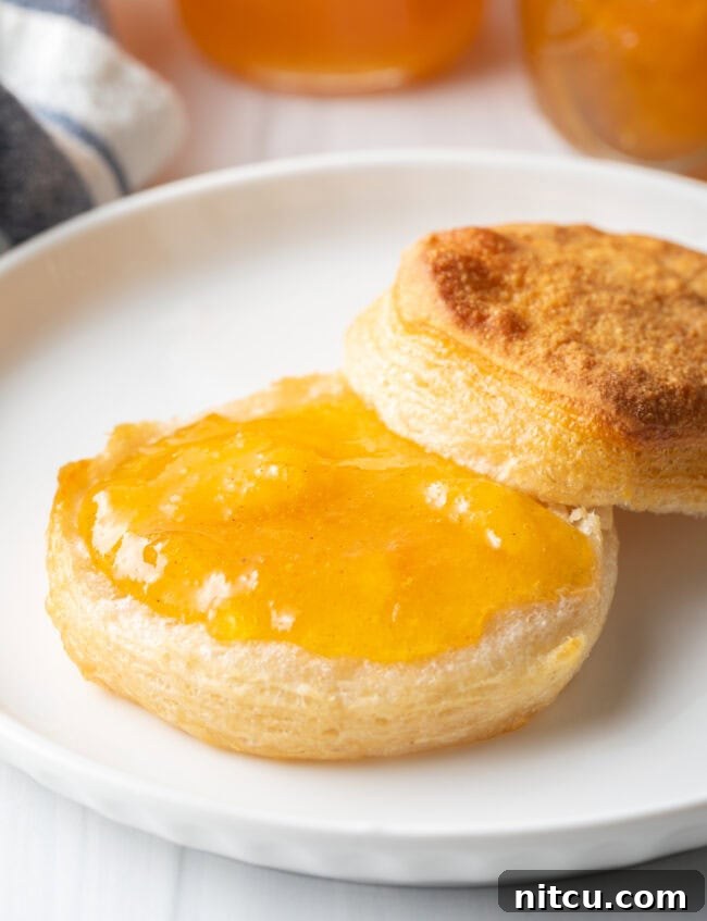 Biscuit split in half on a white plate. Bottom half of biscuit is smeared with peach jam.
