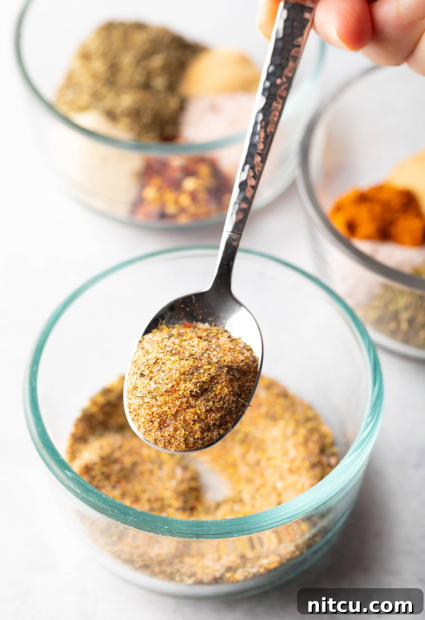 Signature Chicken Seasoning 7 Metal spoon above glass bowl with spice blend. In the spoon is a scoop of seasoning, held to camera.