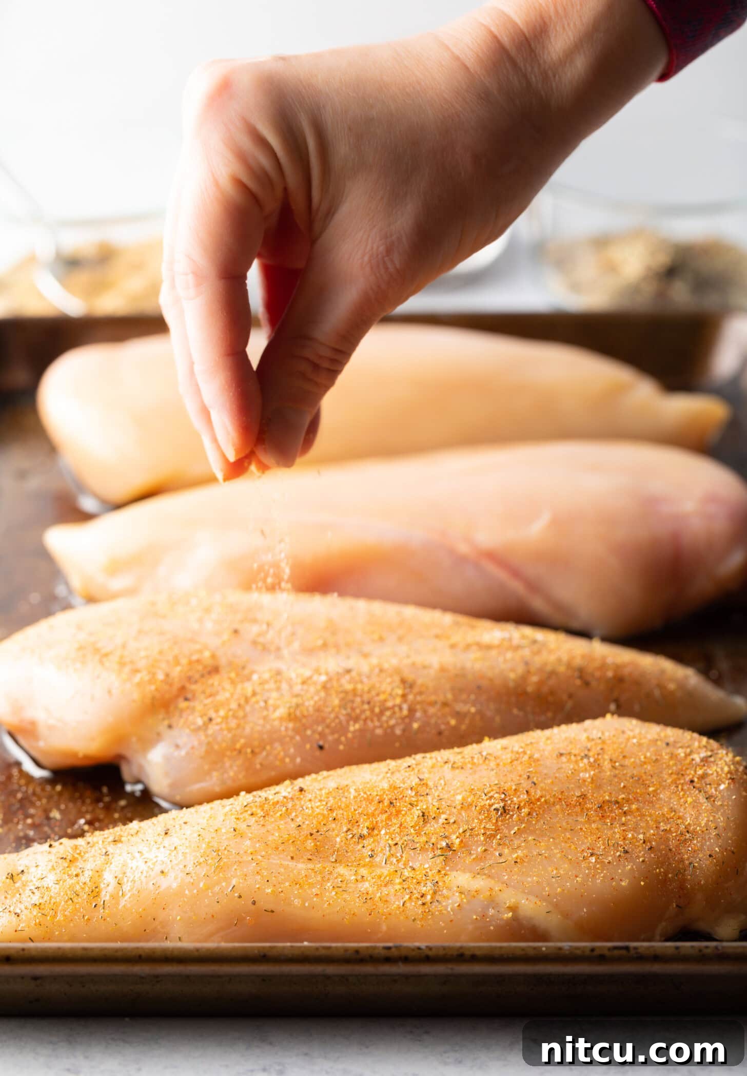 Signature Chicken Seasoning 8 Hand sprinkling seasonings over raw chicken pieces laying out on a metal baking sheet.