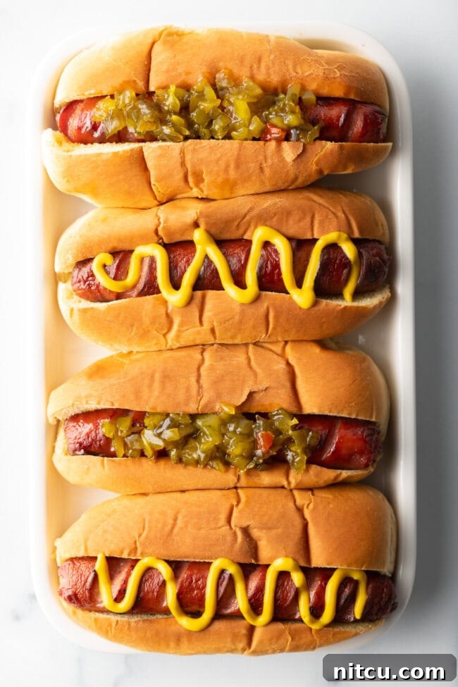 Crispy Air-Fried Franks 4 Top down view. Four hotdogs in buns placed side by side on a white rectangle plate. The toppings alternative between relish and zigzag of mustard.