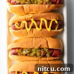 Top down view. Four hotdogs in buns placed side by side on a white rectangle plate. The toppings alternative between relish and zigzag of mustard.
