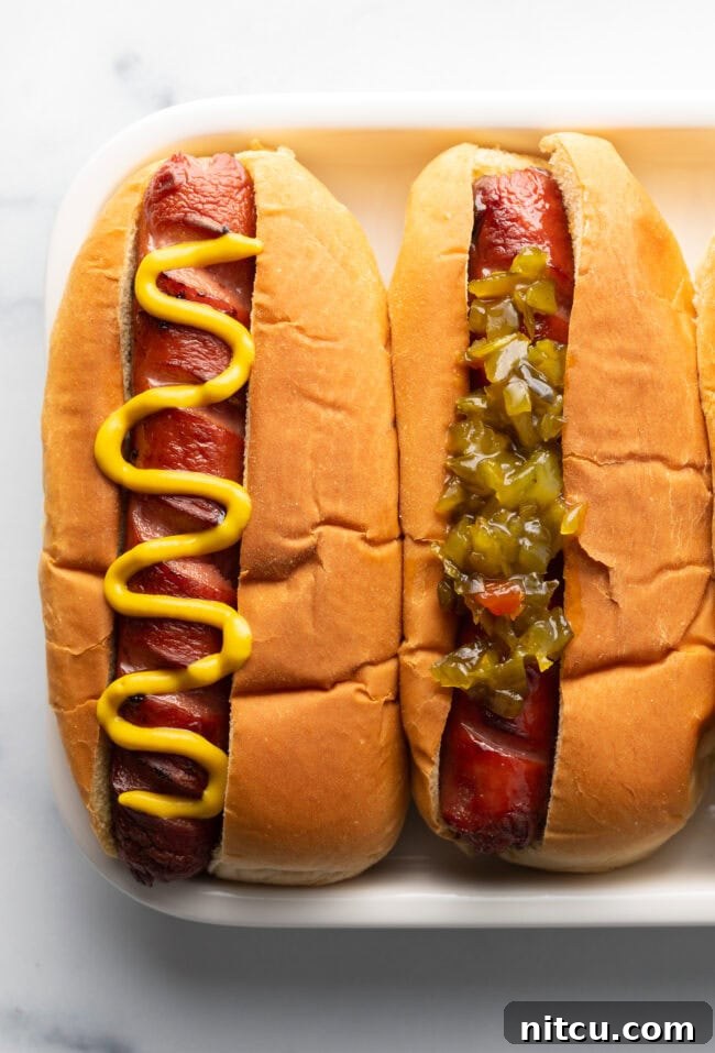 Crispy Air-Fried Franks 10 A top down view of two hot dogs on a white plate. One hot dog has zig zag mustard and the other one has relish.