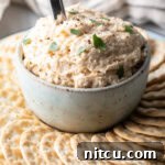 Off-white bowl of smoked fish dip on the center of a round serving plate, with circular crackers all around. A metal spoon is sticking out of the dip.