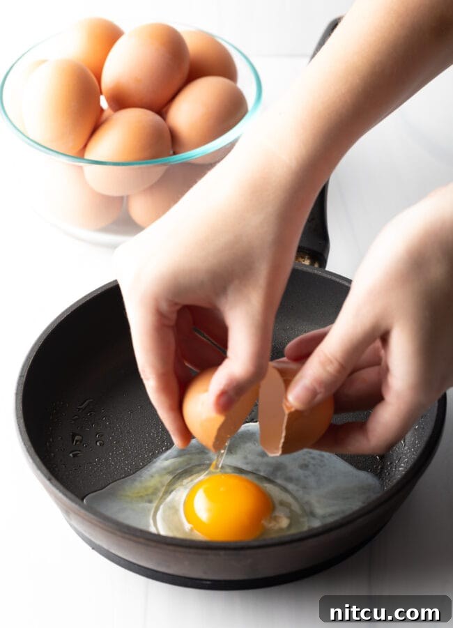 Mastering Over Easy Eggs 7 Hands cracking an egg just above a black frying pan.