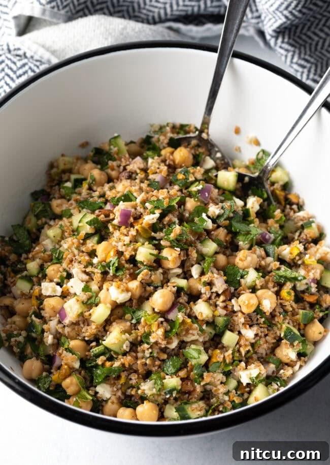 Jennifer Aniston's Go-To Bulgur Salad 2 Large white bowl filled with bulgur wheat, veggies, and feta salad. Two metal salad spoons are in the bowl as well.