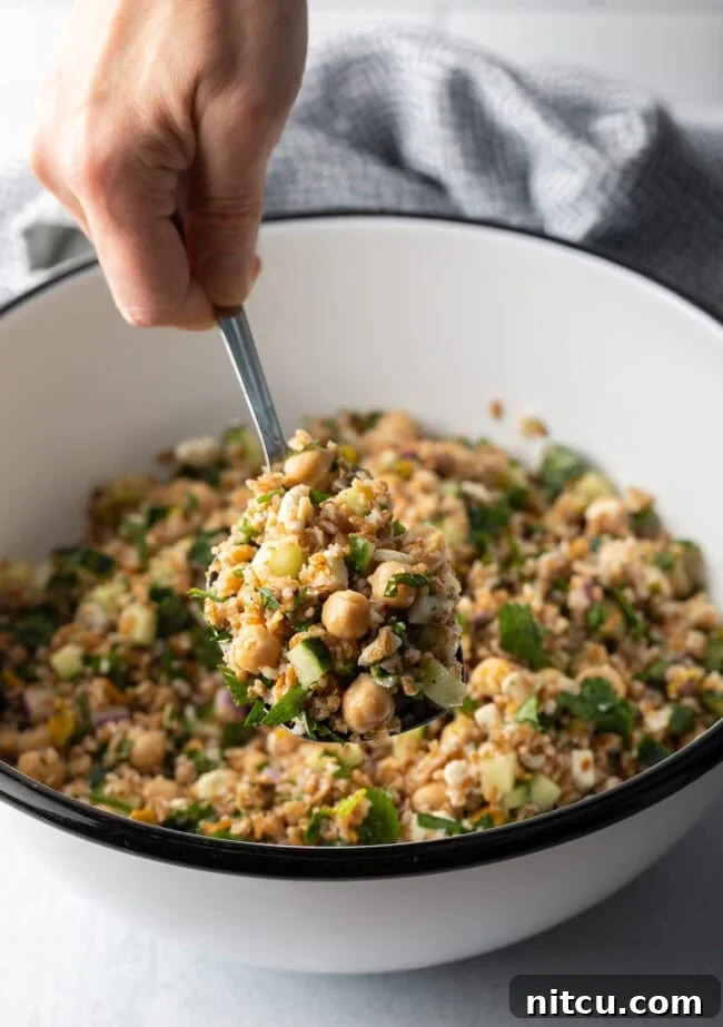 Jennifer Aniston's Go-To Bulgur Salad 13 Hand with metal spoon scooping a serving of salad from a white bowl filled with bulgar wheat, veggies, and feta salad.