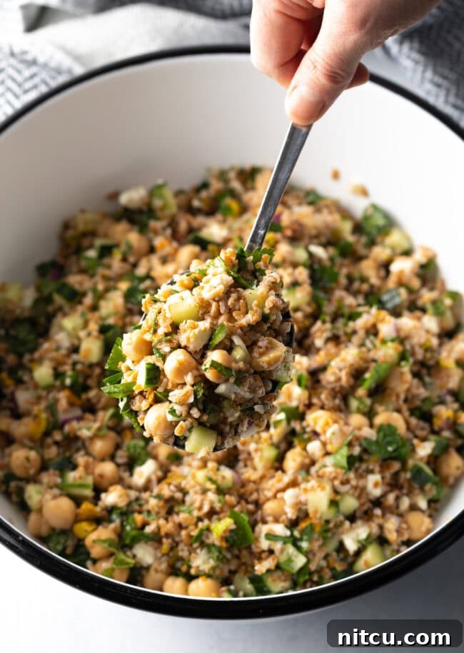 Jennifer Aniston's Go-To Bulgur Salad 5 Hand with metal spoon scooping a serving of salad from a white bowl filled with bulgar wheat, veggies, and feta salad.