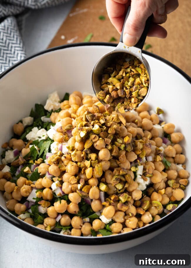 Jennifer Aniston's Go-To Bulgur Salad 9 Hand with metal measuring cup, adding pistachios to the bowl.