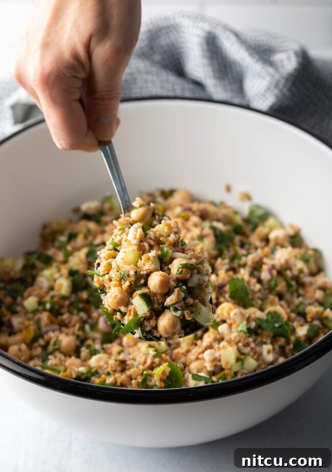 Jennifer Aniston's Go-To Bulgur Salad 10 Hand with metal spoon scooping a serving of salad from a white bowl filled with bulgur wheat, veggies, and feta salad.