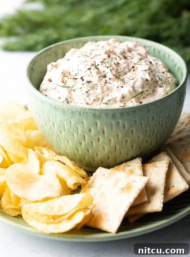 Irresistible Clam Dip 2 A vibrant green bowl filled with creamy clam dip, beautifully arranged on a white plate and surrounded by an assortment of crispy potato chips and crackers.