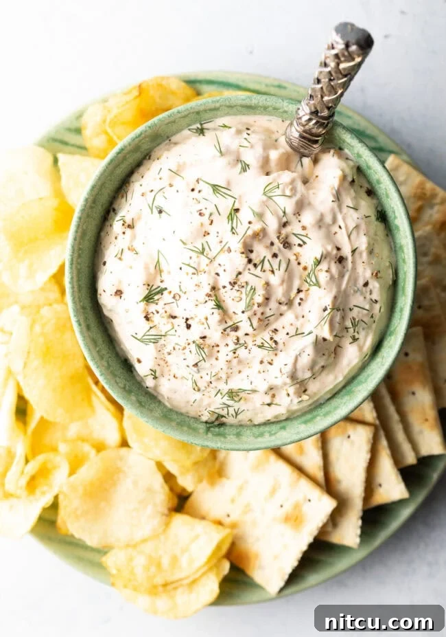 Irresistible Clam Dip 3 A top-down view captures a green bowl overflowing with creamy clam dip, carefully placed on a white plate. It is encircled by an inviting spread of various potato chips and crisp crackers, ready for dipping.