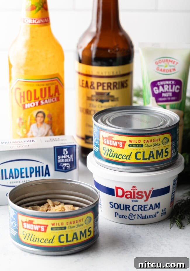 Irresistible Clam Dip 4 A perfectly arranged shot showcasing all the essential recipe ingredients. Featured are a bottle of Cholula hot sauce, a bottle of Worcestershire sauce, two cans of minced clams, a block of cream cheese, and a container of sour cream, all waiting to be transformed.