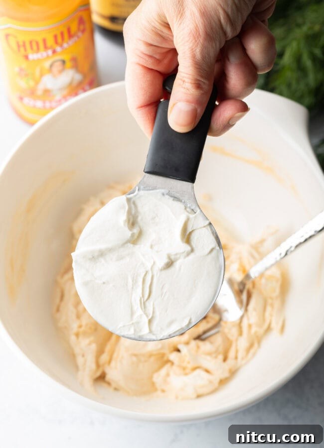Irresistible Clam Dip 5 A hand holds a measuring cup, pouring creamy sour cream into a white bowl already containing a seasoned cream cheese mixture, signifying the next step in the recipe preparation.