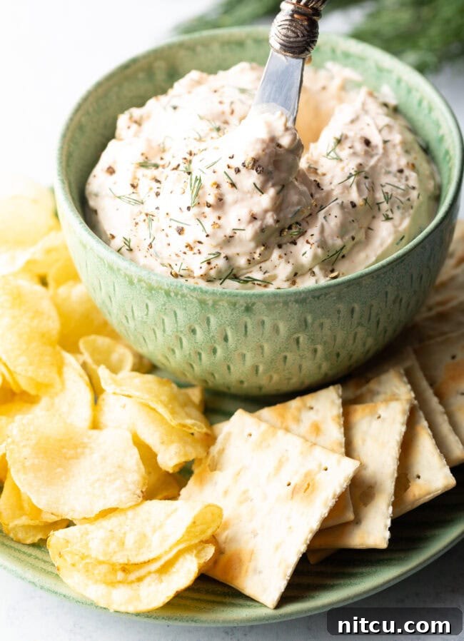 Irresistible Clam Dip 7 A beautifully presented green bowl brimming with creamy clam dip, sitting on a pristine white plate, encircled by a generous selection of potato chips and crackers.
