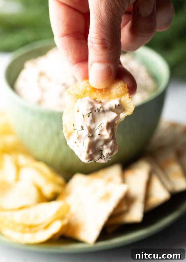 Irresistible Clam Dip 8 A hand skillfully scoops a generous portion of homemade clam dip onto a crisp potato chip, holding it up towards the camera in an inviting gesture.