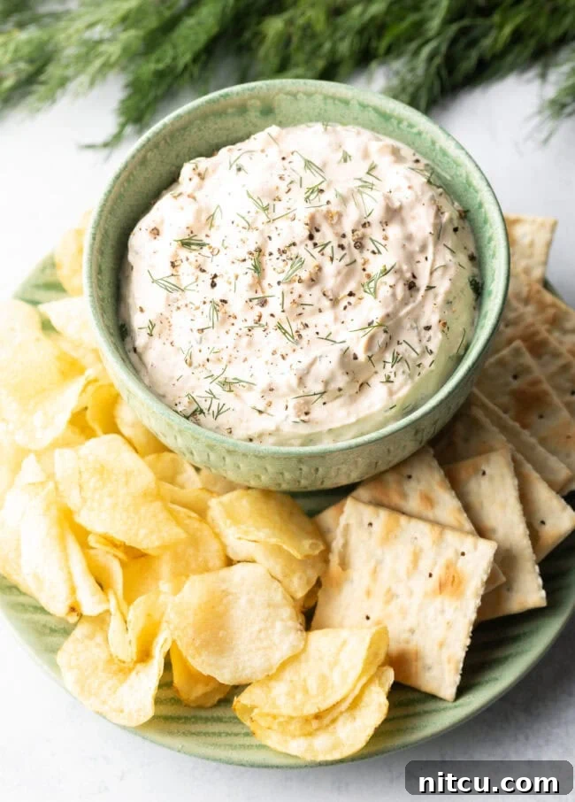 Irresistible Clam Dip 9 A top-down perspective highlights a inviting green bowl, generously filled with the best clam dip recipe, artfully placed on a white plate and encircled by a variety of crispy potato chips and crackers.