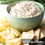 Green bowl filled with creamy clam dip, set on a white plate surrounded by potato chips and crackers.
