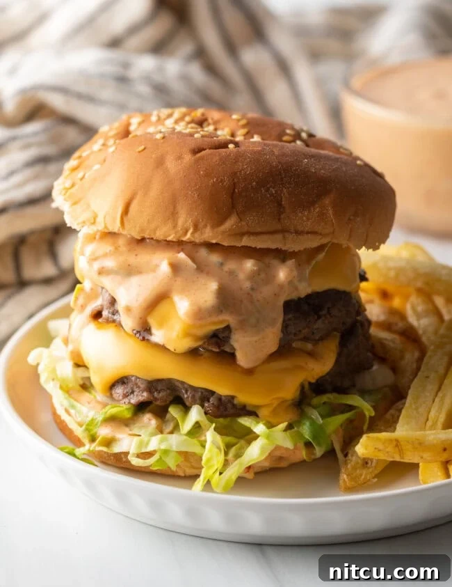 Double patty stacked cheeseburger with homemade Big Mac sauce next to a serving of french fries on a white plate, ready to be enjoyed.