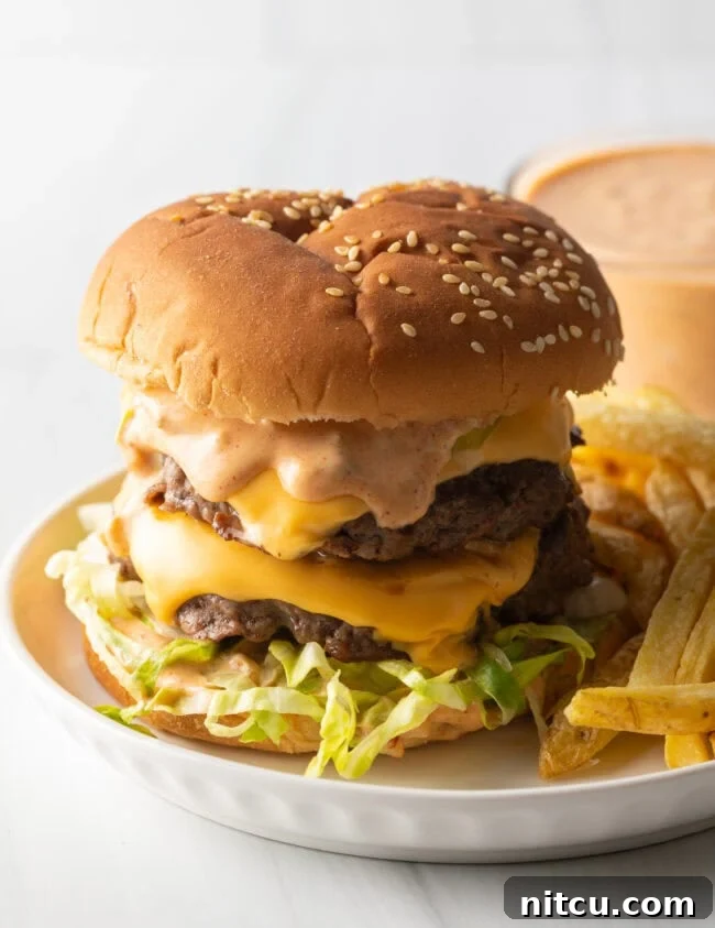 A visually appealing double patty stacked cheeseburger with homemade Big Mac sauce, accompanied by a side of crispy french fries on a white plate, ready to be served.